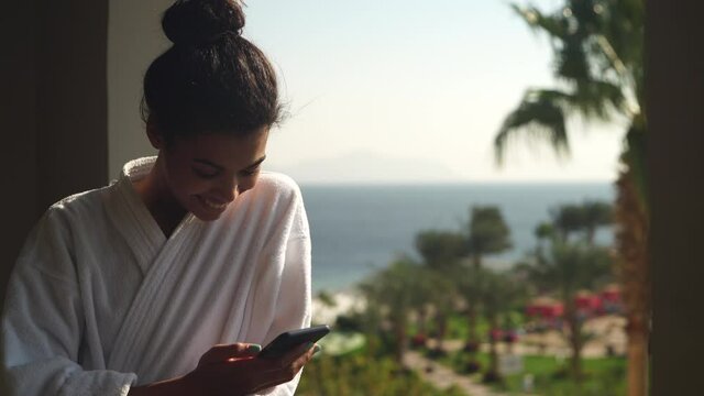 Sharing moment. Young woman in bathrobe chatting, taking pictures of beautiful tropical landscape and sending them to friends, standing on hotel balcony. Vacation, technology concept