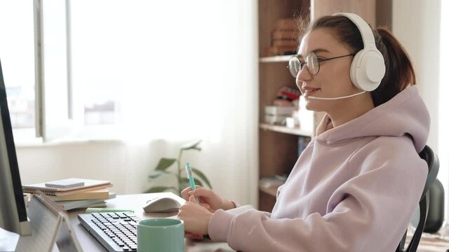 Attractive woman in headphones and glasses is studying with teacher online by video call, preparing for exam