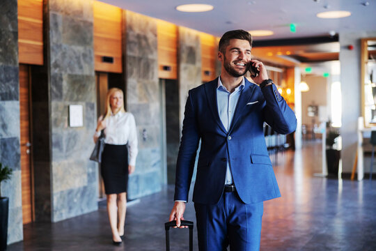 Smiling Middle-aged Man Dressed In Formal Wear Walking In The Lobby Of A Fancy Hotel And Talking On The Phone. He Is Pulling His Suitcase.