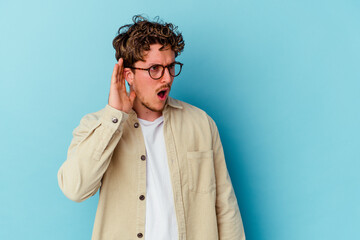 Young caucasian man wearing eyeglasses isolated on blue background trying to listening a gossip.