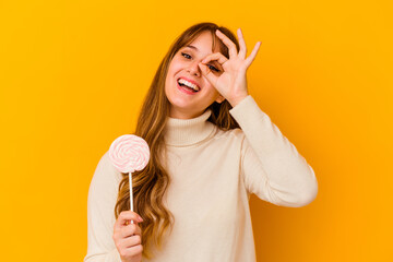Young caucasian woman holding a lollipop isolated on yellow background excited keeping ok gesture on eye.