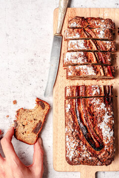 Whole grain banana bread with wallnuts and dried dates, sliced and served on a wooden cutting board with a hand grabbing one piece and a knife.