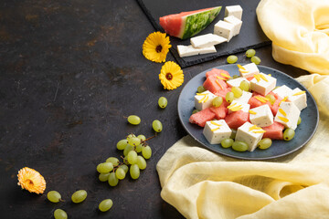 Vegetarian salad with watermelon, feta cheese, and grapes on blue ceramic plate on black concrete background. Side view, copy space.