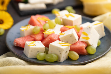 Vegetarian salad with watermelon, feta cheese, and grapes on blue ceramic plate on black concrete background. Side view, selective focus.