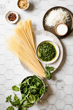 Dry linguini on a plate with a bowl of basil pesto, next to a bowls of fresh spinach, grated Parmesan cheese, crushed red pepper and toasted breadcrumbs.
