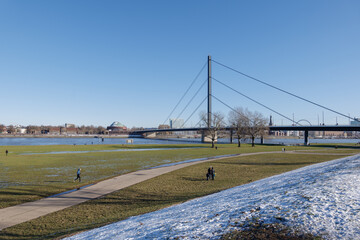 Fototapeta na wymiar Outdoor sunny view of natural riverside of Rhine River covered with slush snow and ice pond with people do winter recreation and background cityscape of Düsseldorf, Germany in winter season.