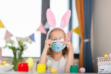 Pretty little girl wearing bunny ears and disposable medical face mask and painting easter eggs at home during coronavirus covid-19 outbreak. 