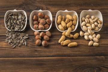Sunflower seeds, peanuts, hazelnuts, and pistachios spill out of cups on a wooden table.