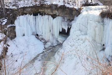 Waterfall frozen in winter with huge icicles hanging off rocks and water starting to flow down the...