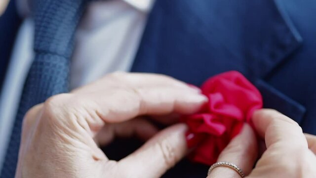 The caring hands of a woman putting a lapel flower (a floral decoration) on the elegant suit of the groom. Handheld closeup shot.

