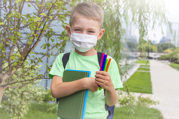 New normal, back to school. Schoolboy wearing medical mask and backpack holding textbook and felt-tip pens outdoors. Coronavirus protection. Keep your distance