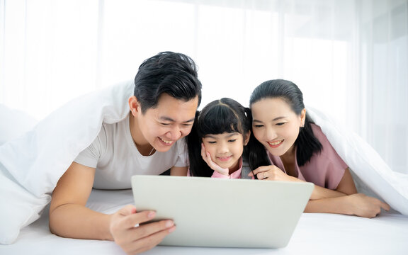 Happy asian family people leisure in bedroom together. Father and mother with daughter relaxing and using laptop on bed - Powered by Adobe