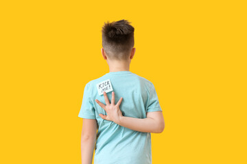 Sticky note with text KICK ME on back of little boy against color background. April Fools Day prank