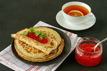 Traditional Russian Crepes Blini stacked in a plate with red caviar and hot lemon tea on wooden background. Maslenitsa traditional Russian festival meal. Russian food, russian kitchen.