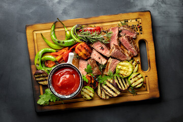 Fototapeta na wymiar Sliced fried medium steak with grilled vegetables and ketchup sauce on wooden cutting board on dark background top view