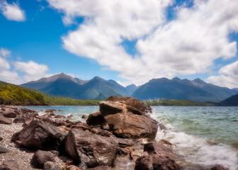 Fototapeta na wymiar Waves crashing on the rocks at Lake Manapouri on a beautiful day with blue skies and puffy clouds. Lake Manapouri is the gateway for the remote fjord of Doubtful Sound in New Zealand, South Island.