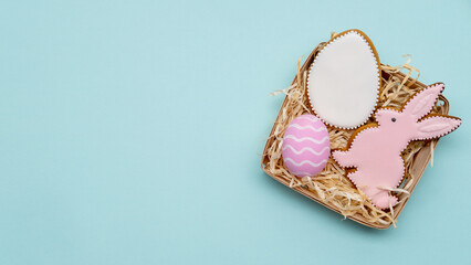 Easter gift. Holiday food ornament. Homemade pastry decoration. Sweet pink white gingerbread bunny biscuit painted egg set for girl in rustic basket with straw isolated on blue copy space background.