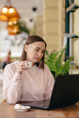 Woman drinks coffee and works at a laptop while sitting in a cafe