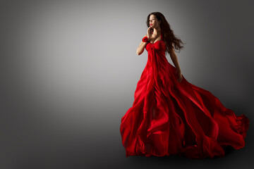 Fashion Woman in Red Dress. Beauty Model dancing in Long Evening Gown fluttering on Wind Black Hair flying in Air over Gray Background