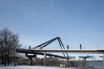 Obraz premium Outdoor sunny view of Fussgängerbrücke, pedestrian bridge, at Rheinpark Bilk and field covered by snow on promenade of Rhine river in winter season in Düsseldorf, Germany. 