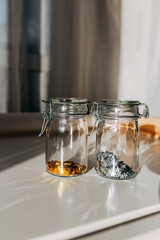 Golden and silver sparkles in glass jar. Gold glitter under sun. Two cans with glitters in shape of heart and stars on white table.