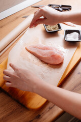 Raw chicken breast on a wooden board, near salt, spices and a knife. A woman prepares chicken breast in the home kitchen.