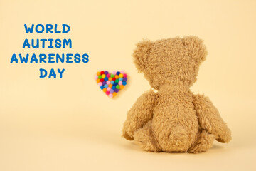 World Autism Awareness Day concept - sad teddy bear and multicolored defocused heart against yellow background. Autism spectrum disorder and child mental health concept. Selective focus