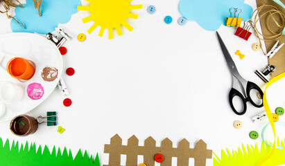 Fototapeta premium The background is a view from above on a white table with children's creativity. On the table of colored paper the sun, clouds, grass, fence, palette, scissors, colored buttons.