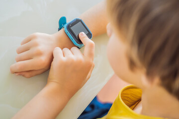 Little boy sitting at the table and looking smart watch. Smart watch for baby safety. The child makes school lessons, listening to music, calling friends