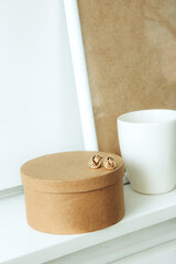 Still life. Cup of coffee, blank beige poster frame, craft round box and beautiful gold earrings on the white wall. Side view