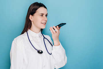 Smiling female doctor wearing white medical uniform and stethoscope holding phone speak activate virtual digital voice assistant on smartphone, consult patient online, isolated on blue background