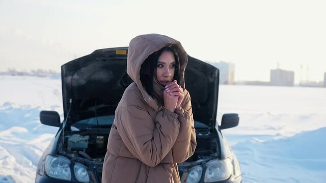 Sad frozen woman in warm jacket stands near broken automobile with open bonnet on empty snowy road in countryside in cold winter evening