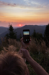 Mobile phone in arms of young man on landscape background. Tourist taking picture of beautiful sunrise in mountain. Hands with smart phone. A man takes a photo for memory at dawn.