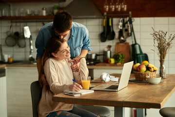 Affectionate man kissing his girlfriend who is working on laptop at home.