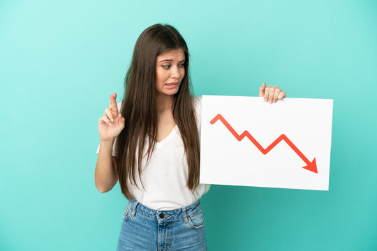 Young caucasian woman isolated on blue background holding a sign with a decreasing statistics arrow symbol with fingers crossing