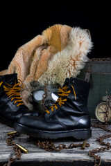 Leather boots with rusted chains and bullet shells with military gas mask and fur coat in background resting on wooden table