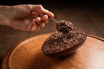 Hand sticking a metallic spoon in a stuffed chocolate easter egg with grated chocolate on the top on a wooden stand on a wooden table. 