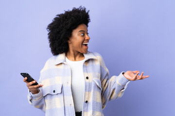 Young African American woman using mobile phone isolated on purple background with surprise facial...