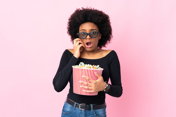 Young African American woman isolated on pink background with 3d glasses and holding a big bucket of popcorns