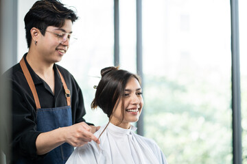 Professional male stylist cutting woman's hair in salon. The man hairdresser using scissors cut the young Asian girl sitting in the shop looking at mirror with smile. Beauty salon business concept.