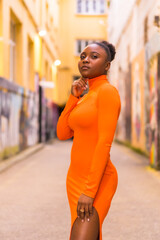 Fashionable urban style with a black African girl in an orange dress and black heels on a city street. Portrait looking at camera