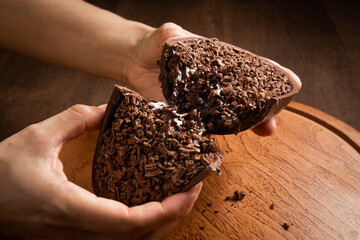 Woman hands crack a stuffed chocolate easter egg with grated chocolate on the top on a wooden stand...