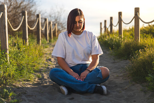 joven pelirroja y con piercings reflexionando mientras realiza yoga en un peque&ntilde;o camino en un paseo vestida con vaqueros y camisa blanca