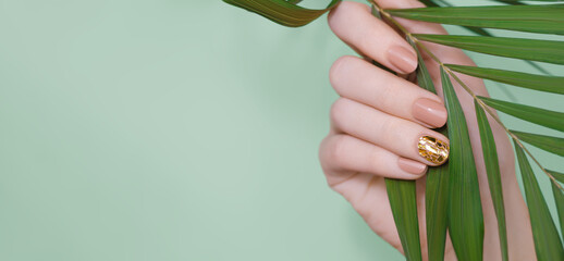 Female hand with beige nail design. Beige nail polish manicure and glitter golden nail art. Female hand hold tropic leaf - obrazy, fototapety, plakaty