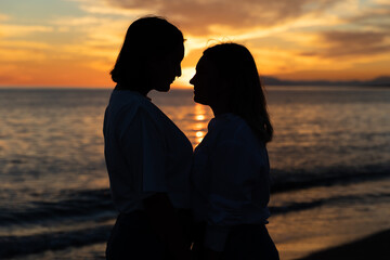 Joven pareja lesbiana mirándose bajo un atardecer a través de una silueta oscura con precioso mar...