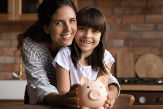 Portrait of happy Latino young mom and small 8s daughter recommend smart living and saving. Smiling Hispanic mother and little teen girl child hold piggybank feel economical making investment.