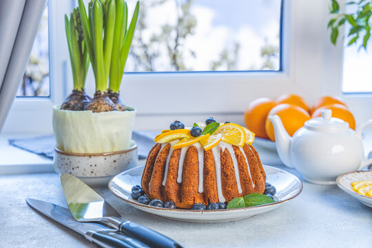 Orange bundt cake with blueberry surrounded fruits, plant and cutlery on light table near window. Family breakfast concept