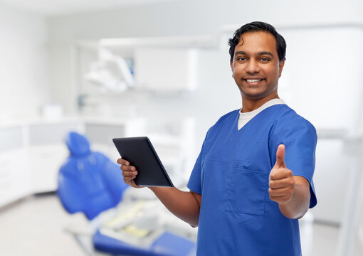 medicine, dentistry and technology concept - happy smiling indian doctor or male dentist in blue uniform with tablet pc computer showing thumbs up over dental clinic background