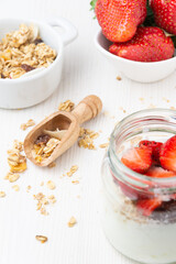 Top view of jar with yogurt with muesli and strawberries, spoon, bowl with muesli and another with strawberries, selective focus, on white table, vertical