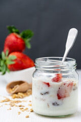 Close-up of jar of yogurt with strawberries and muesli with spoon, bowl with strawberries, selective focus, on white table, gray background, vertical